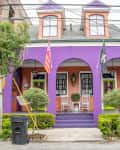 Purple house with green doors, rocking chairs on porch, and flags. Flanked by trees and shrubs on a city street.