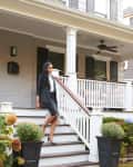 Woman in business attire walking down steps of a large porch with white railings and potted plants.