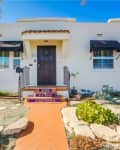 White stucco house with black awnings, colorful tiled steps, and a landscaped front yard with palm trees.