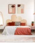 Modern bedroom with a scalloped headboard, abstract art, and plants. White dresser, round mirror, and coral bedding.