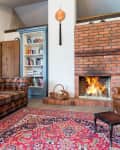 Cozy living room with plaid sofa, leather armchair, brick fireplace, bookshelf, and red patterned rug.