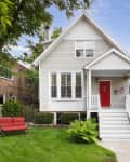 White house with red door, front porch, and American flag, surrounded by green lawn and trees, with a red bench.