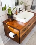 Wooden bathroom vanity with white sink, black faucet, potted plant, and toiletries on a tiled wall background.
