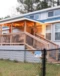 Cozy house with a blue metal roof, wooden porch, and decorative lights, surrounded by a fenced yard and greenery.