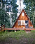 A-frame cabin with wooden exterior, surrounded by tall trees, and a small porch with steps leading to the entrance.