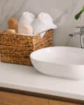 A modern bathroom countertop with a white oval sink, chrome faucet, woven basket of towels, and a potted plant.