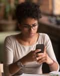 Person with glasses and curly hair looking at a smartphone, sitting at a table with papers and a plant in the background.
