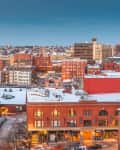 Snow-covered rooftops and brick buildings in a cityscape at dusk, with a view of distant water.
