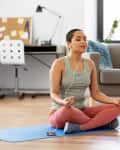 Woman meditating on a blue yoga mat in a living room, wearing headphones, with a sofa and desk in the background.