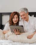 Couple in bed smiling at a tablet, with a lamp and books on a bedside table.
