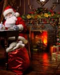 Santa Claus reading a letter at a wooden desk, with a gift box, near a decorated Christmas tree and a lit fireplace.