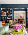Cozy living room with green sofas, colorful pillows, a marble coffee table, and a bookshelf filled with books.