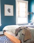 Teal bedroom with wooden bed, layered bedding, abstract wall art, and patterned rug.