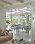 Living room with pastel sofa, floral pillows, and a shell chair, leading to a dining area with green chairs and large windows.