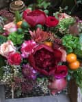 Vibrant floral arrangement with red peonies, pink roses, green foliage, and small orange fruits on a rustic wooden chair.