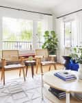Living room with wooden chairs, round table, books, and plants by large windows with white curtains.