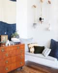Mid-century dresser with books and plants beside a cozy nook with patterned cushions and hanging ceramic decor.