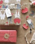 Gift wrapping paper with paper doll cutouts, red and brown patterns, scissors, and twine on a craft table.