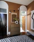 Brown room with a wooden dresser, mirror, modern light fixture, and a patterned rug. Open door reveals greenery outside.