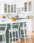 White kitchen with green island, white bar stools, glass cabinets, and a bowl of oranges on the counter.