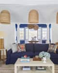 Living room with navy sofa, colorful pillows, wicker lamps, and a view into a dining area with blue accents and large windows.