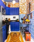 Colorful kitchen with blue cabinets, abstract wallpaper, orange tiger rug, and stainless steel countertop with fruit and decor.