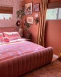 Pink bedroom with patterned wallpaper, velvet bed, abstract bedding, wall art, plants, and gold decor accents.