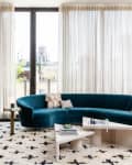 Modern living room with teal curved sofa, gray armchairs, geometric coffee tables, and large windows with sheer curtains.
