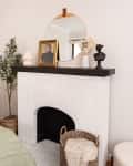White brick fireplace with black mantel, round mirror, framed portrait, vase with flowers, and a bust sculpture.