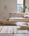Minimalist living room with a wooden daybed, beige cushions, textured rug, and a potted plant by the window.