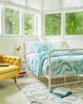 Bright bedroom with a yellow armchair, white metal bed, teal patterned bedding, and large windows overlooking greenery.