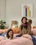 Woman and two children on a bed with pink bedding, surrounded by plants and abstract art on the wall.