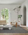 Beige sectional sofa and armchair in a living room with arched window, abstract art, and potted plants on a checkered rug.