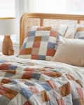 Patchwork quilt and pillows in blue, orange, and white on a bed with a wooden headboard, next to a ceramic lamp.