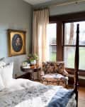 Bedroom corner with floral armchair, round wooden table, vase of flowers, and portrait painting near large window.