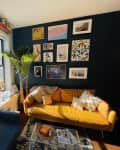 Cozy living room with a yellow sofa, blue couch, gallery wall, and a tall plant by a window with a lamp and books.