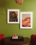 Green wall with two framed portraits above a round table, red chairs, books, and glasses, under a woven pendant light.