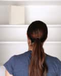 Person with brown ponytail looking into an empty white cabinet with a single box on the shelf.