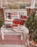 Porch with Christmas decor, plaid blanket, pillows, small trees, and a rustic table with mugs and festive items.