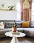 White living room with gray sectional sofa, pink drapery, and boho accents like plants and a cane chair and woven light fixture
