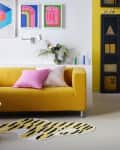 yellow black and white squiggly shaped rug with line striped tiger like pattern, yellow couch, pink and pink and white striped pillow, black wood chair, black glass book case, colorful graphic art, white floating shelf behind couch, plants, colorful splat patterned rug