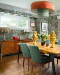 Wooden dining table with teal chairs in room with patterned wallpaper and short curtains.