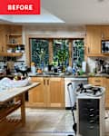 Cluttered kitchen with wooden cabinets, a large window, various kitchen appliances, and a table with utensils and containers.