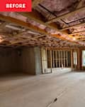 Open space under construction with exposed wooden beams, pink insulation, and concrete flooring. Two large windows and a door lead outside.