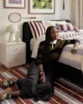 A woman in a striped sweater sits on a colorful rug beside a bed with patterned bedding, a lamp, and a tennis racket on the wall.