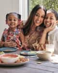 Woman with two children at a table with snacks, including carrots, pretzels, and dips, near a window with greenery outside.