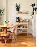 view into dining room with arched doorway wood round table with wood chairs, wood floors, cream walls, kitchen island with coffee maker, toaster oven, floating shelf above with plant, teapot, coffee accessories