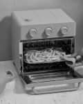 Person placing fries in a small oven on a kitchen counter with utensils and a bowl of fruit.