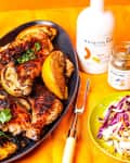 Grilled chicken with lemon wedges on a black platter, Brightland olive oil bottle, and a plate of shredded cabbage.