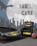 Batman-themed kids' room with car bed, armchair, cityscape mural, and "Sam's Cave" sign.